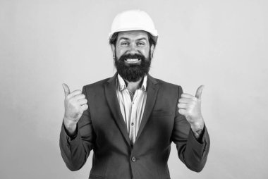 Sakallı mutlu adam sert şapka takıyor. Mühendislik kavramı. Mimari ve bina. Erkek müteahhit koruma için resmi takım elbise ve kask giyiyor. İnşaat projesinde çalışıyorum. Başparmağını kaldır. inceleniyor
