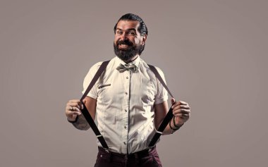 Olgun bir adam pantolon askısı takar ve papyon takar. Saçları, sakalı, modası ve berberi çok bakımlıdır.