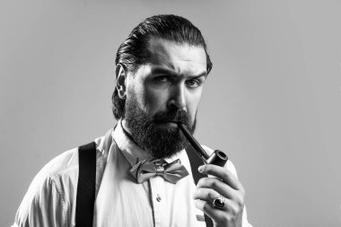 Erkeklik ve karizma. resmi erkek tiryaki. Askılı ve papyonlu eski moda sakallı hippi pipo içiyor. Kendine güvenen ve klasik giyinmiş zarif bir adam.
