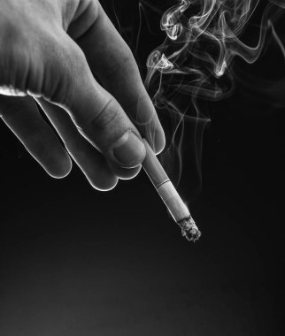 Male hand hold cigarette butt with smoke swirls dark background, copy space, nicotine addiction