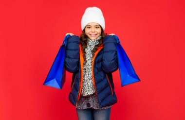 Şişkin ceketli ve şapkalı mutlu çocuk. Kırmızı arka plandan alışveriş yapan genç kız..