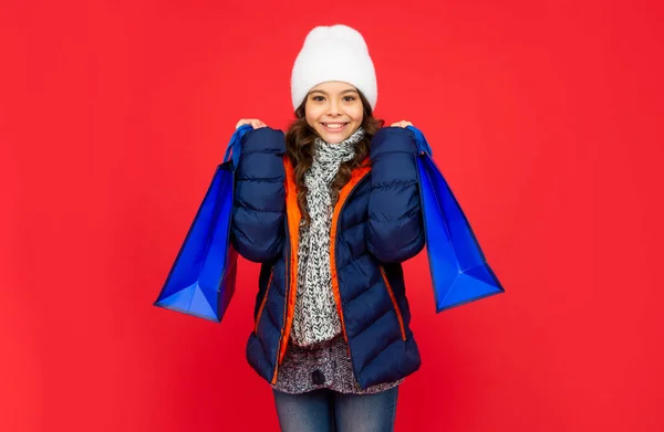 Um miúdo feliz de casaco e chapéu. menina adolescente depois de fazer compras no fundo vermelho. — Fotografia de Stock