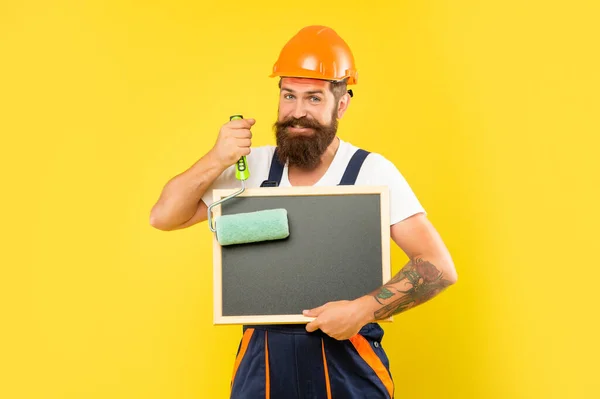 Positive Mann Renovierer in Helm und Arbeitskleidung halten Farbroller und Tafel — Stockfoto