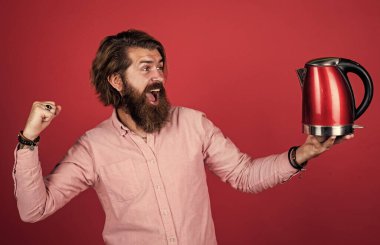 Gömlekli sakallı adam elektrikli su ısıtıcısı tutuyor. Sıradan bir adam modern teknolojiyi günlük hayatta kullanır. Bıyıklı mutlu hipster, kaynar su. Erkek ev idaresi. Çay içme konsepti. yüksek kalite