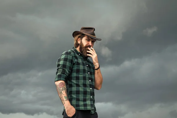 Hipster maduro com barba em chapéu de cowboy. Um hipster caucasiano brutal com bigode a fumar cigarro. Mau hábito masculino. tipo ocidental confiante em camisa quadriculada. homem barbudo — Fotografia de Stock