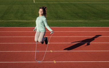 Antrenman rutini. Spor giyimli bir çocuk ip atlarken üzerine atlıyor. Çocuk yarış pistinde egzersiz yapıyor. sağlıklı çocukluk. Açık havada egzersiz yapmak için. İp atlama. Genç kız stadyumda ısınıyor.