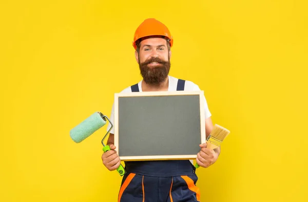 Glücklicher Mann mit Helm und Arbeitskleidung hält Pinsel und Tafel in der Hand — Stockfoto