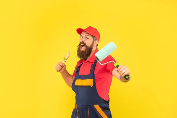 Hombre barbudo enojado en ropa de trabajo mantenga rodillo de pintura y cepillo sobre fondo amarillo — Foto de Stock