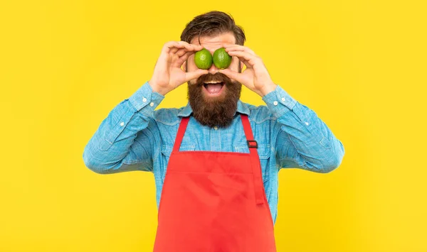 Meglepett férfi kötényben gazdaság friss lime szemek sárga háttér, gyümölcs eladó — Stock Fotó