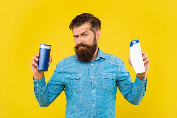 Жестокий бородатый человек выбрать бутылку кондиционера для волос на желтом фоне, уход за волосами — стоковое фото