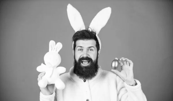 Easter when you are parent. Man handsome face wear white bunny ears. Easter bunny. White bunny symbol of easter. Guy with toy and easter egg on violet background. Enjoy tenderness. Spring holiday