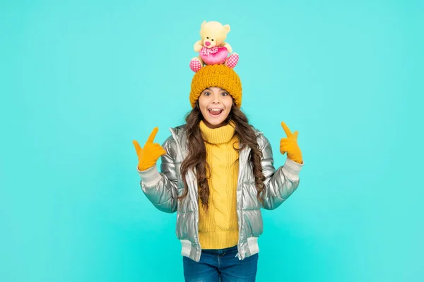 Verbaasd kind in hoed en handschoenen houden speelgoed punt vinger op blauwe achtergrond, cadeau — Stockfoto