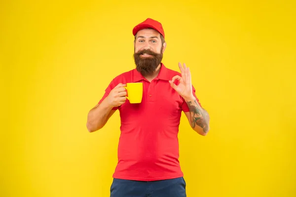 Felice uomo barbuto in casual berretto rosso e maglietta dando OK tenendo tazza sfondo giallo, caffè — Foto Stock