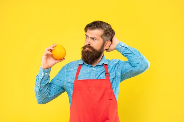 Μπερδεμένος άνθρωπος με ποδιά κοιτάζοντας φρέσκα φρούτα πορτοκάλι ξύνοντας το κεφάλι κίτρινο φόντο, φρούτων — Φωτογραφία Αρχείου