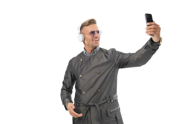Schöner Typ in lässigem Trenchcoat und Kopfhörer lächelt zum Handy, Selfie — Stockfoto