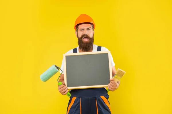 Šťastný muž malíř v helmě a pracovní oblečení držet barvu váleček štětec a tabule — Stock fotografie