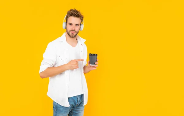 Чоловік у білій сорочці носить навушники точковий палець на чашці кави на жовтому фоні, випийте — стокове фото