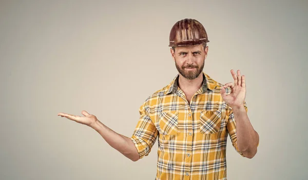 Homem bonito mecânico em capacete de segurança de construção e camisa quadriculada no local de construção apresentando produto com gesto ok, espaço de cópia, qualidade — Fotografia de Stock