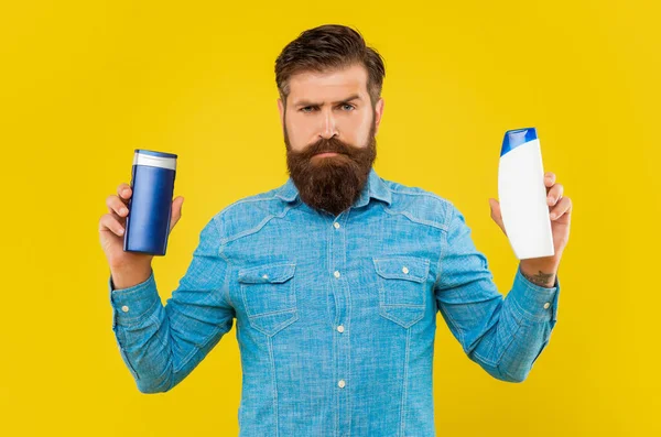 Жестокий бородатый человек выбирает бутылку шампуня на желтом фоне, представляя туалетные принадлежности — стоковое фото