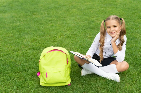 Criança feliz em uniforme com saco escolar leitura livro sentado na grama verde — Fotografia de Stock