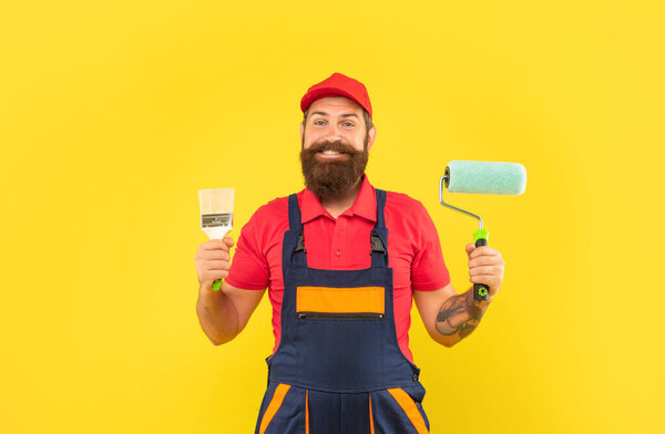 happy bearded man renovator in work clothes hold paint roller and brush on yellow background