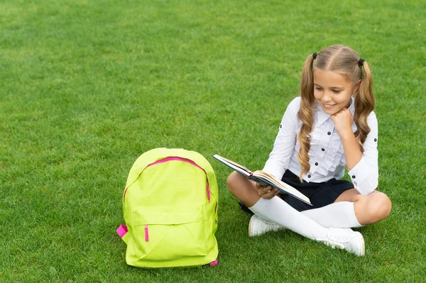 Menina feliz em uniforme com saco escolar leitura livro sentado na grama verde — Fotografia de Stock
