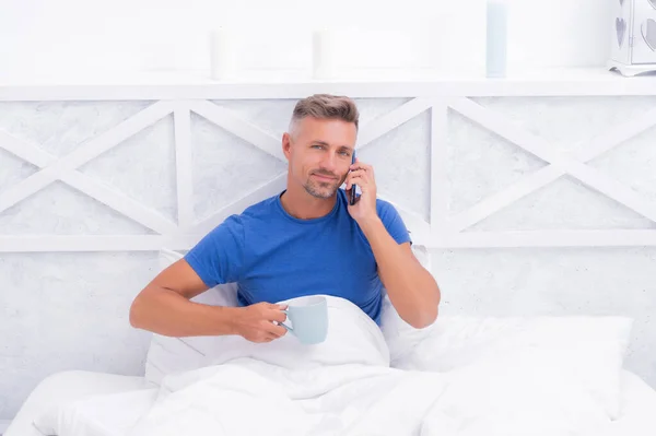Glad kille pratar på mobiltelefon medan du dricker morgonte eller kaffe i sängen, ring — Stockfoto