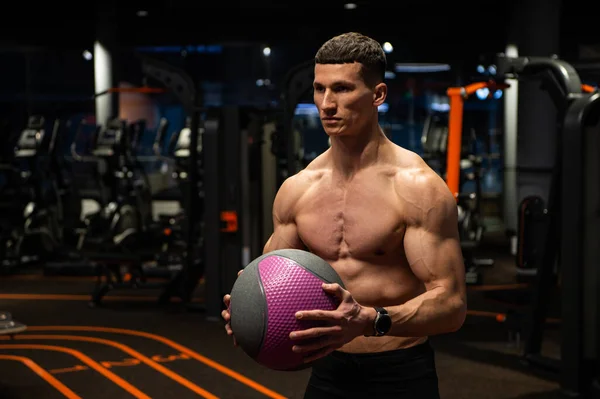 М'язистий чоловік з сорочкою без підгонки торс тримає медичний м'яч для балістичних вправ у тренажерному залі, силові тренування — стокове фото