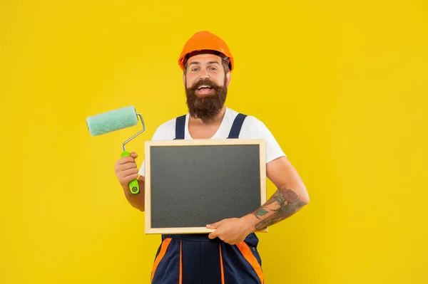Veselý muž malíř v helmě a pracovní oblečení držet barvu váleček a tabule — Stock fotografie