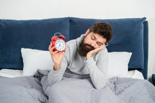 Сонливый мужчина смотрит на будильник, лежащий в постели утром, потеря сна — стоковое фото