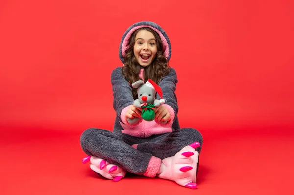 Verbaasd kind in comfortabele pyjama zitten hold xmas speelgoed op rode achtergrond, kerst — Stockfoto