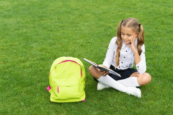 Bambino felice in uniforme con libro di lettura borsa della scuola seduto su erba verde — Foto Stock