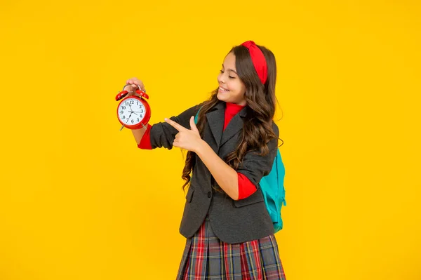 Niño feliz en uniforme con el dedo de la mochila en el reloj despertador sobre fondo amarillo, mañana. — Foto de Stock