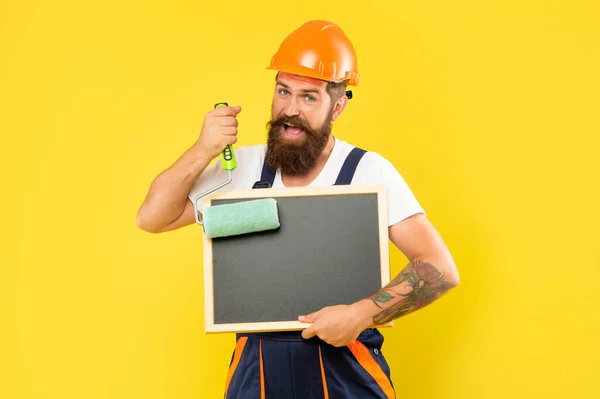 Mosolygós férfi szobafestő sisakban és munkaruhában tartsa festék görgő és táblán — Stock Fotó
