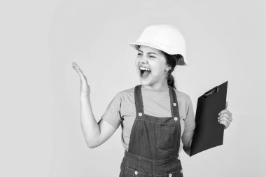 Sert şapkalı kızgın çocuk not alıyor. Çocuk tutulan belge klasörü. Çocuk, güvenlik şapkalı inşaat işçisi olmayı öğreniyor. Kasklı, tabletli küçük çocuk. Onarım planlaması. kızgınlığını ifade et