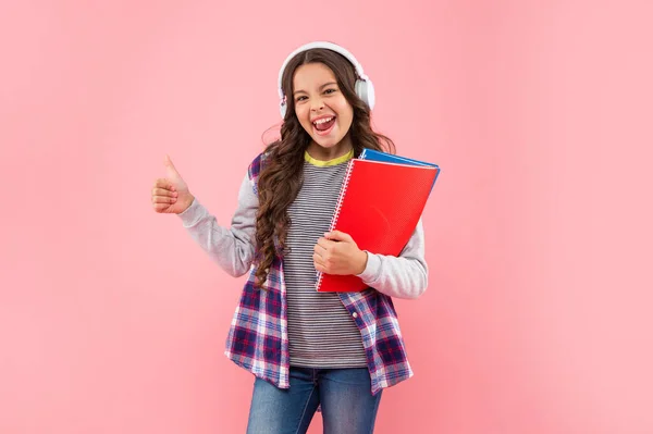 Χαρούμενο παιδί που ακούει μουσική σε ακουστικά με βιβλίο εργασίας σε ροζ φόντο που δείχνει τον αντίχειρα επάνω, γνώση — Φωτογραφία Αρχείου