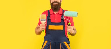 İş kıyafetleri içinde kırpılmış adam dekoratörü. Sarı arka planda boya silindiri ve fırça tutuyor.