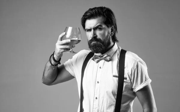 Ваше здоровье. бородатый мужчина в подтяжках пьет виски. жестокий парень-бармен носит бабочку. элегантный бармен. Красивый хипстер пьет стакан рома. Вечеринка с алкоголем. бренди или коньяк — стоковое фото