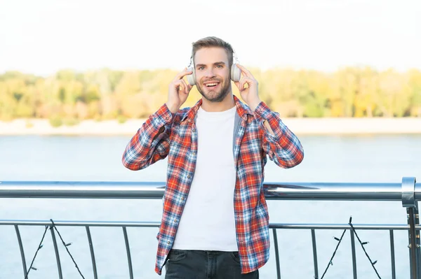 Tıraşsız, mutlu, yakışıklı, kareli gömlekli adam kulaklıkla müzik dinliyor, açık havada müzik dinliyor. — Stok fotoğraf