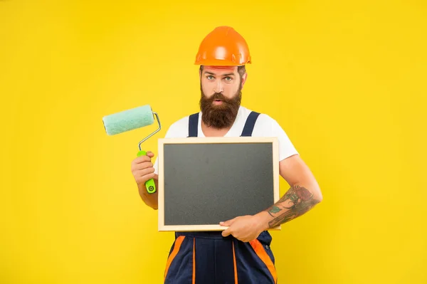 Užaslý muž malíř v helmě a pracovní oblečení držet barvu váleček a tabule — Stock fotografie