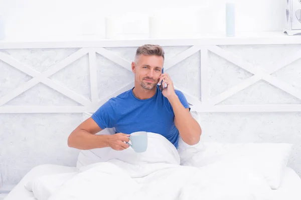Glad man pratar på mobiltelefon medan du dricker morgonte eller kaffe i sängen, ring — Stockfoto