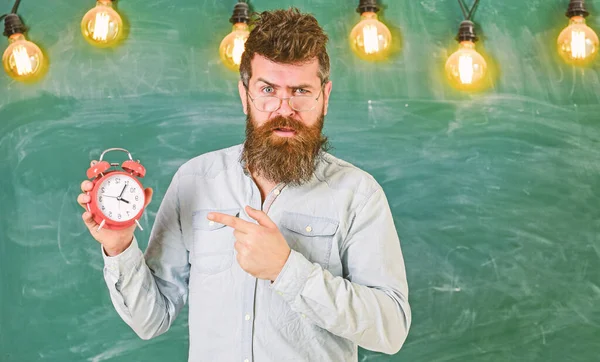 Teacher in eyeglasses holds alarm clock. Lateness concept. Man with beard and mustache on strict face pointing at clock. Bearded hipster holds clock, chalkboard on background, copy space — Stock Photo, Image