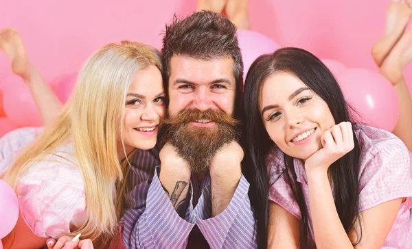 Girls fall in love with bearded macho, pink background. Threesome on smiling faces lay near balloons. Alpha male concept. Man with beard and mustache attracts blonde and brunette girls