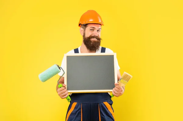 Glücklicher Mann in Helm und Arbeitskleidung hält Pinsel und Tafel mit Kopierraum in der Hand — Stockfoto