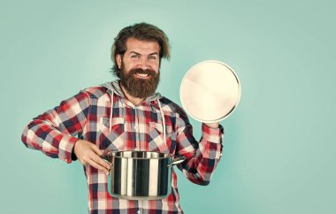 Havalı saç stili olan şık sakallı adam. Kareli gömlek tutacağı takıyor, yemek yapıyor.