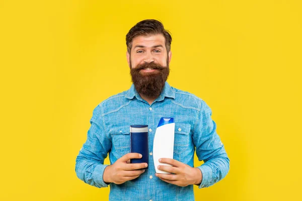 Felice brutale uomo barbuto tenere bottiglia condizionatore di capelli su sfondo giallo, cura dei capelli — Foto Stock