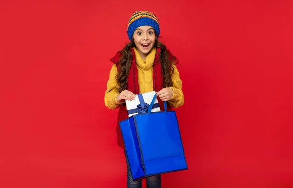 Meghökkent tini lány visel kötött kalap hold bevásárló táska és jelen doboz piros háttér, vásárlás — Stock Fotó