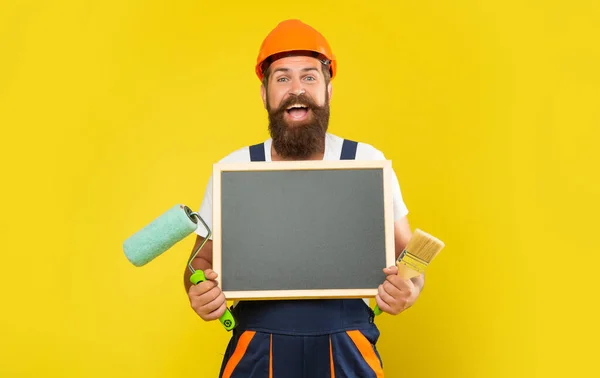 Щасливий чоловік художник у шоломі та робочому одязі тримає пензлик для фарби та дошку з копіювальним простором — стокове фото