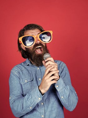 Olgun sakallı adamın uzun saçları var. Parti gözlüğü takıyor ve mikrofonla şarkı söylüyor.