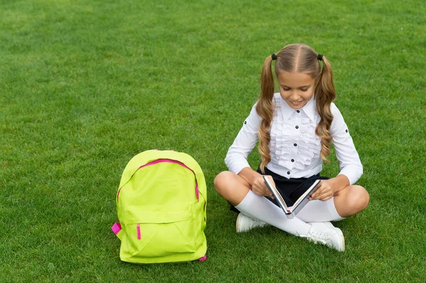 Счастливая девочка с книгой для чтения в школьном портфеле, сидящая на зеленой траве — стоковое фото